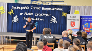 Policjant z mikrofonem oraz umundurowani policjanci siedzący na krzesłach