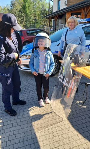 Policjantka oraz dziewczynka na tle policyjnego radiowozu. Na zdjęciu widoczna tarcza z napisem policja oraz osoba biorąca udział w wydarzeniu