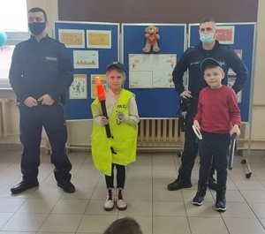 Dwóch policjantów w maseczkach. Dziewczynka w odblaskowej kamizelce z napisem POLICJA trzyma policyjną latarkę, chłopiec pałkę służbową oraz tarczę.